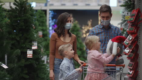 The-family-chooses-the-scenery-in-the-supermarket.-A-happy-family-in-medical-masks-in-the-store-buys-Christmas-decorations-and-gifts-in-slow-motion